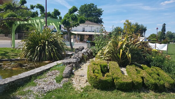 Un jardín bien cuidado con un sendero de piedra, abundante vegetación y un edificio con la leyenda 'Le Vieux Logis' al fondo.