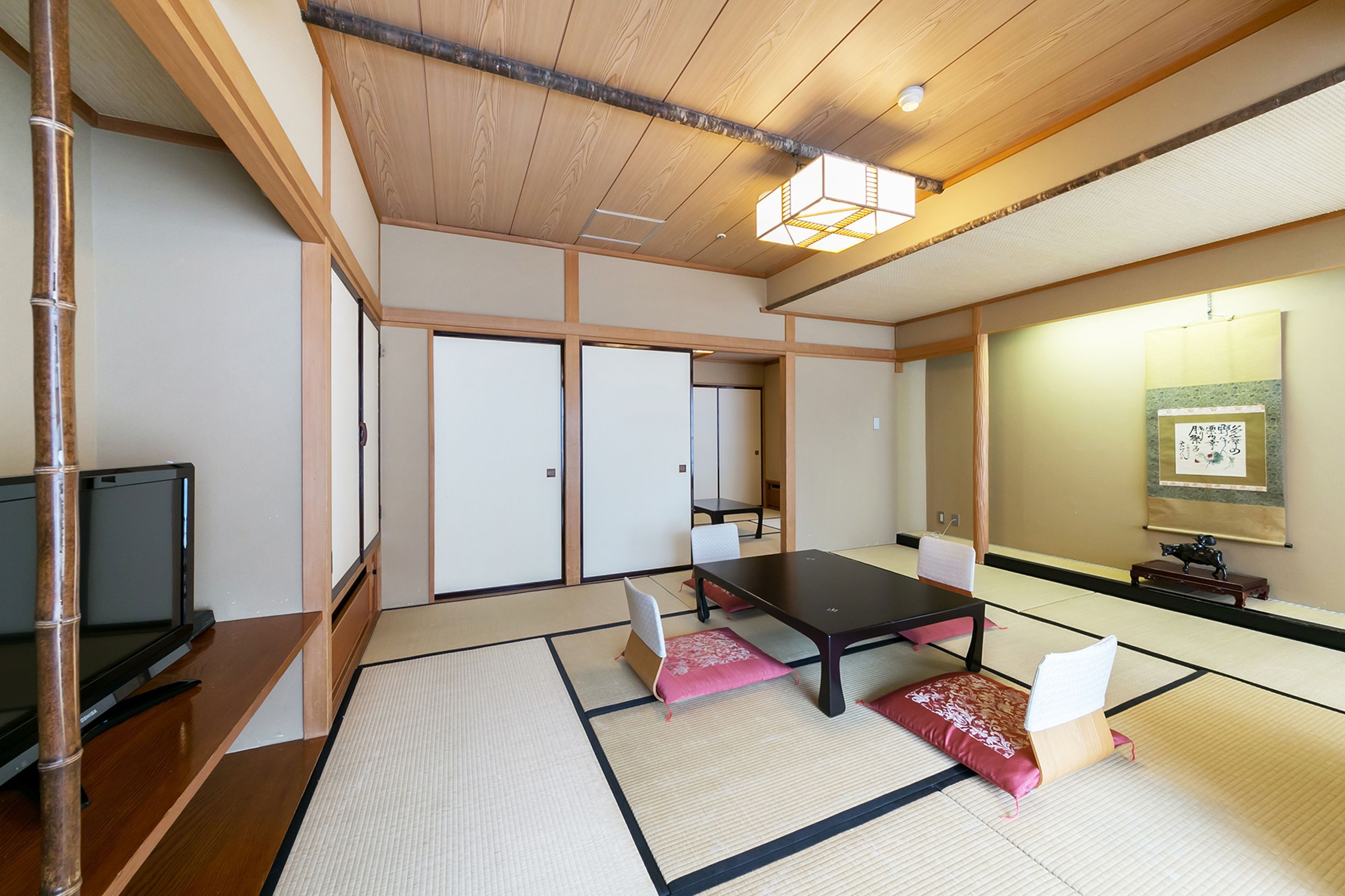 A traditional Japanese room with tatami flooring, a low table, cushions, sliding doors, and a television.