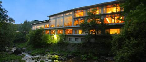 A modern hotel with multiple floors, large windows, and a river in the foreground.