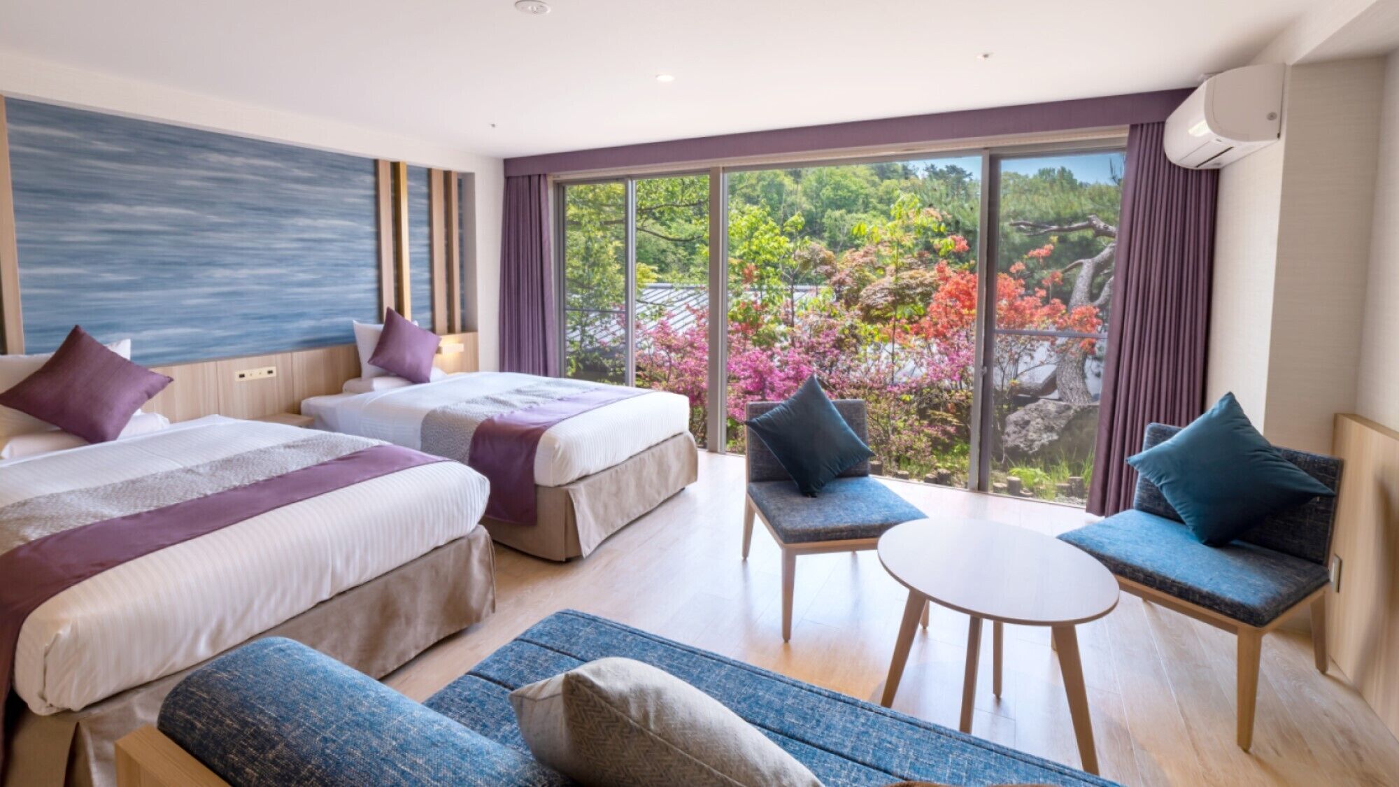 A hotel room with two beds, a sofa, a chair, a small table, and a large window with a view of trees and flowers.