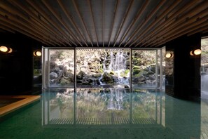 Une piscine intérieure moderne, dotée d’une paroi vitrée, d’un plafond en bois et offrant une vue sur une cascade.