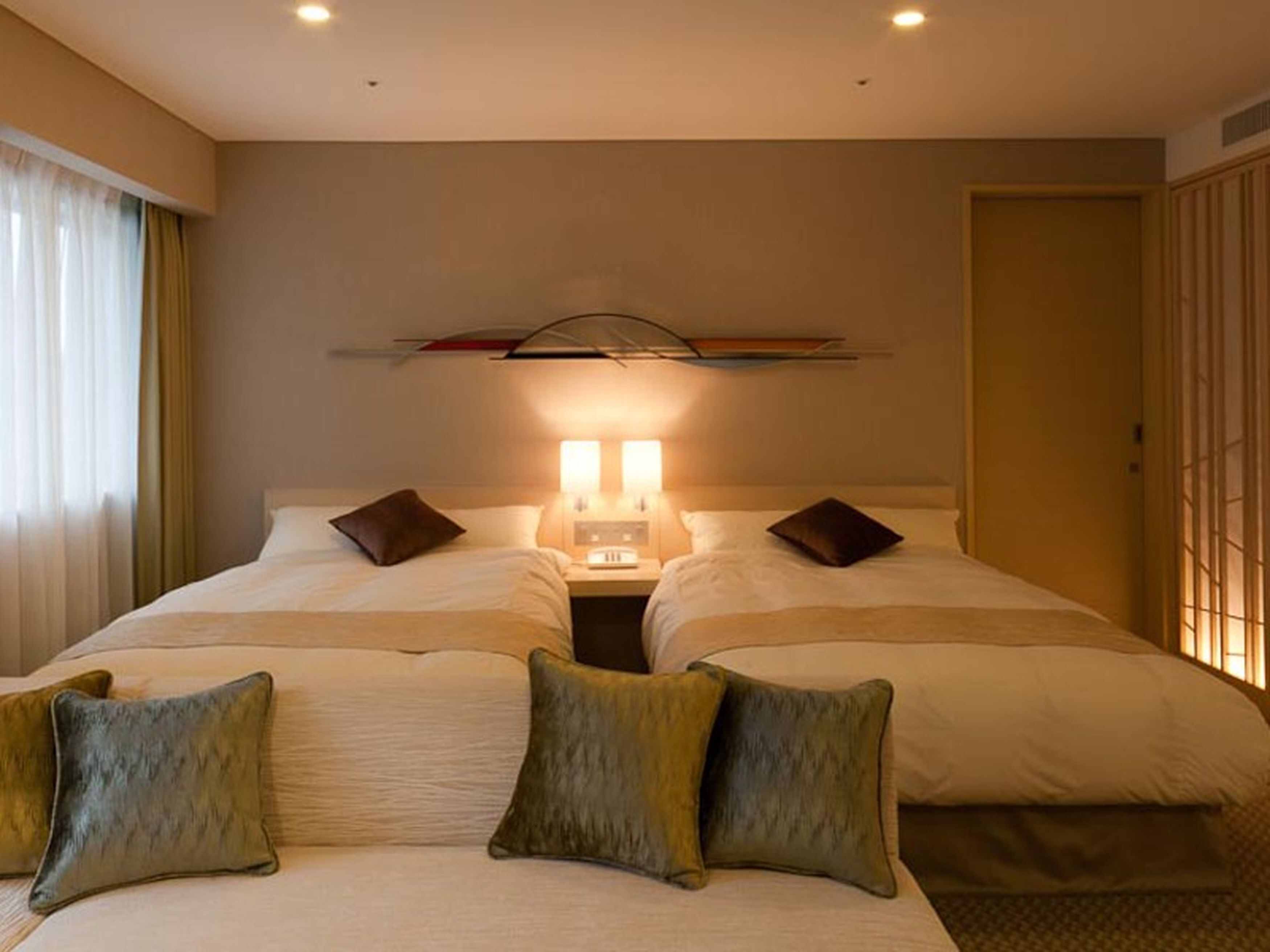 A hotel room with two beds, a nightstand with lamps, and a large window with curtains.