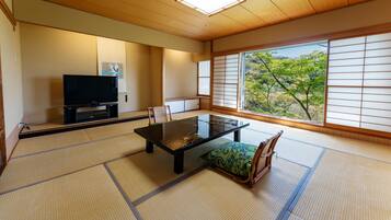 A traditional Japanese room with tatami flooring, a low table, a television, and a view of trees through shoji screens.