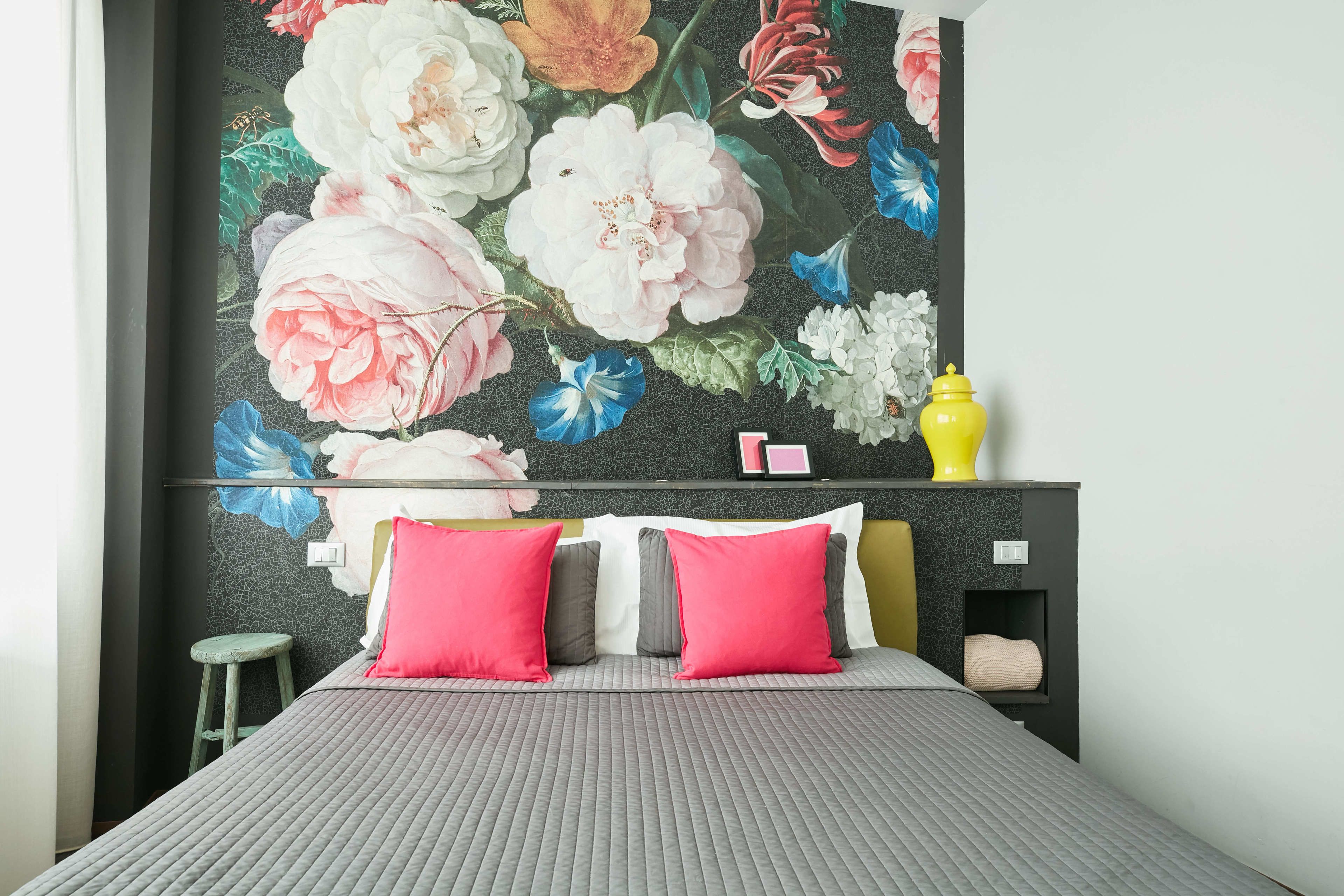 A bedroom with a large floral mural on the wall, a bed with grey bedding and pink pillows, a small stool, and a yellow lamp on a nightstand.