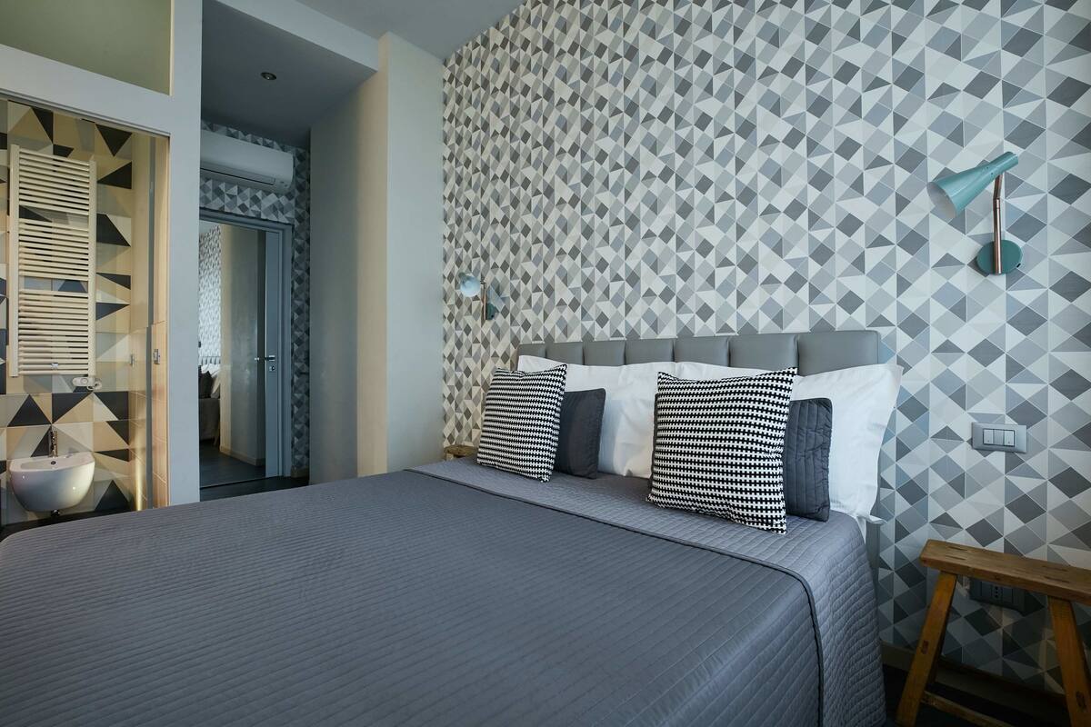 Un dormitorio moderno con pared de diseño geométrico, una cama gris con muchas almohadas, una mesita de noche y un taburete de madera.