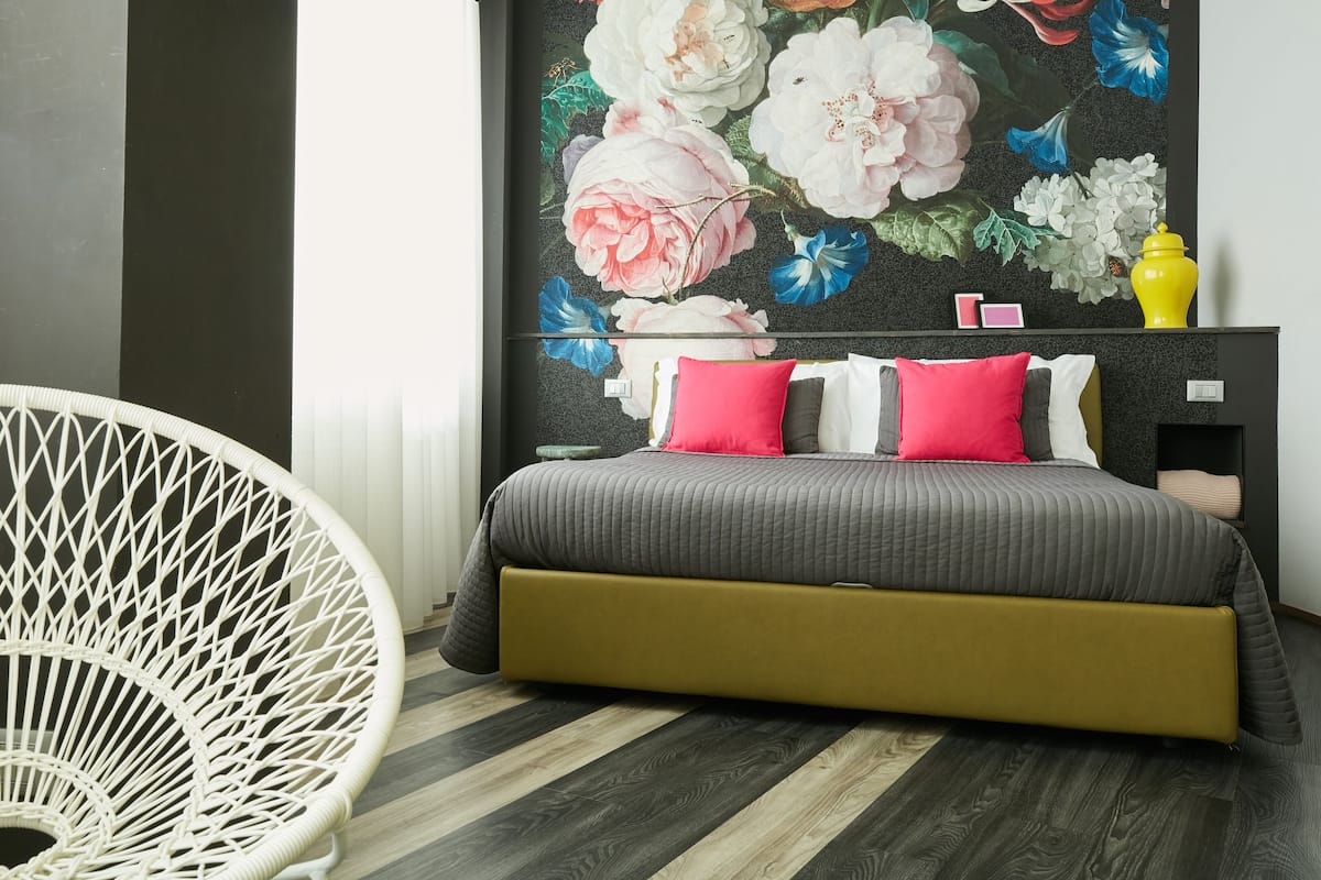 Un dormitorio con una cama, una silla blanca, un mural floral y un jarrón amarillo.