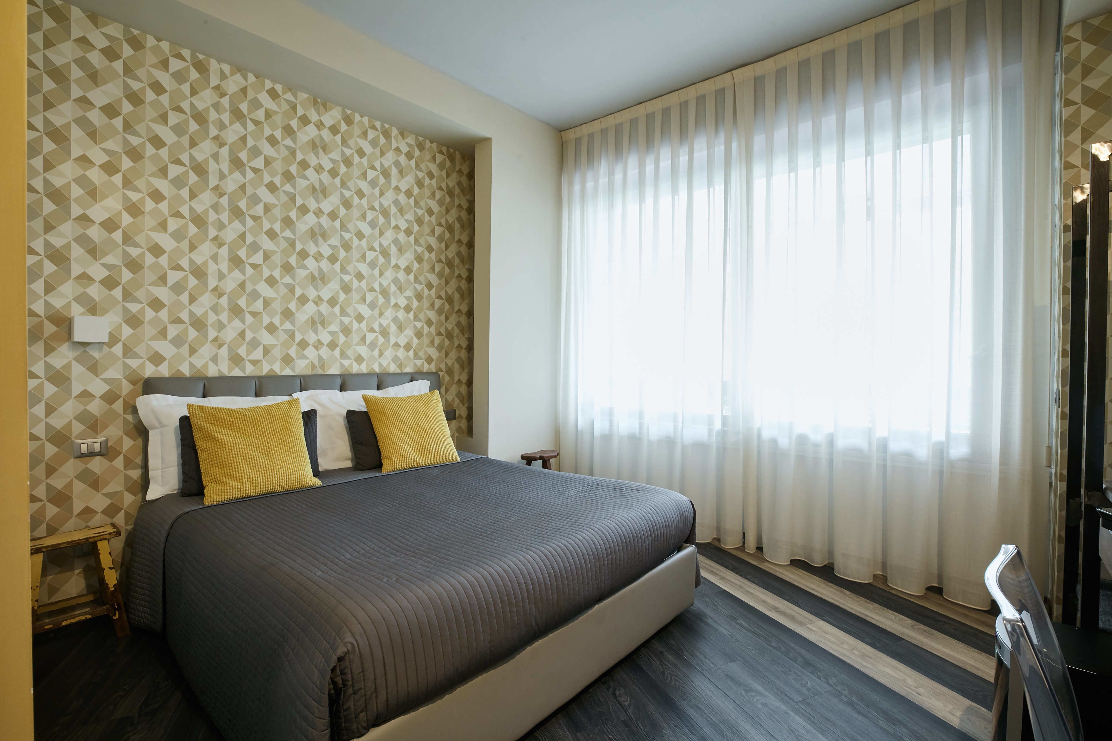 Una habitación de hotel moderna con una cama grande, papel tapiz de estampado geométrico, suelo de madera y un ventanal con cortinas translúcidas.