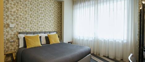 Una habitación de hotel moderna con una cama grande, papel tapiz de estampado geométrico, suelo de madera y un ventanal con cortinas translúcidas.