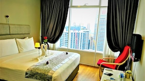 A hotel room with a large bed, a bedside table with a lamp, a red chair, and a view of the cityscape through the window. - Saba Suites at Vortex KLCC Bukit Bintang (Kuala Lumpur)