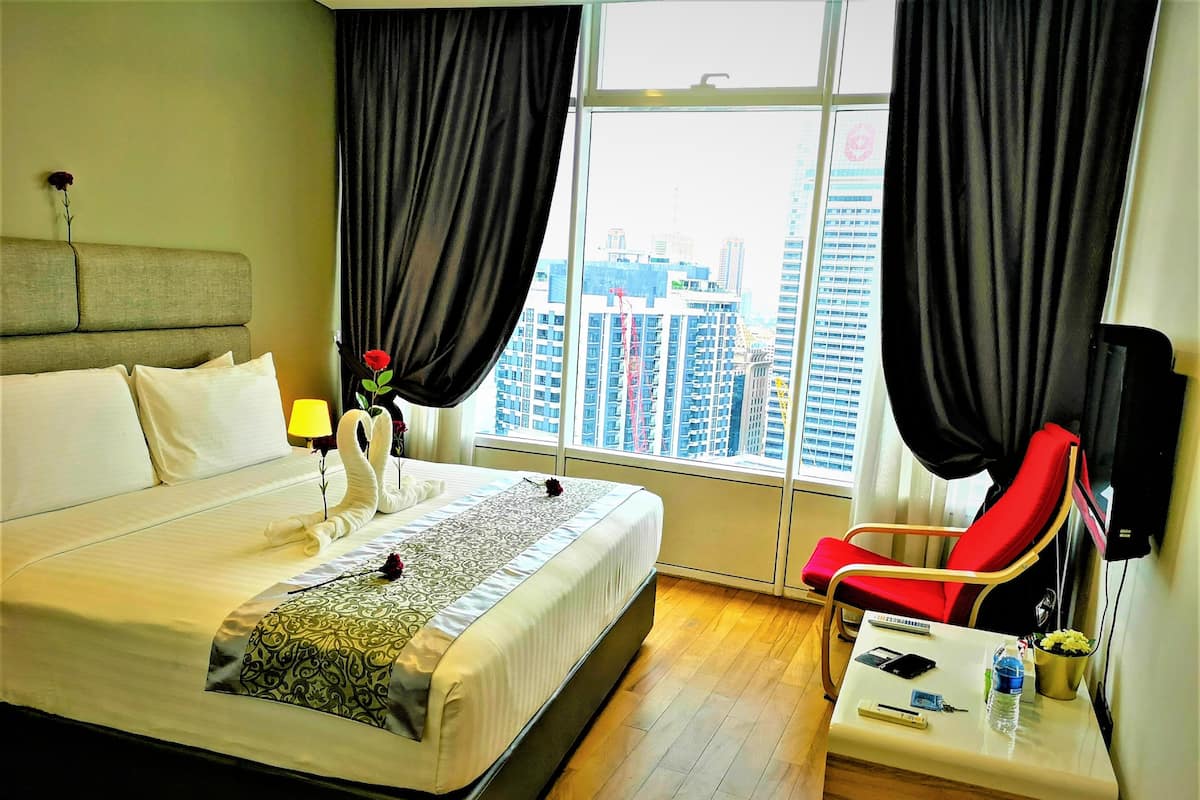 A hotel room with a large bed, a bedside table with a lamp, a red chair, and a view of the cityscape through the window.