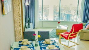 A modern living room with a dining area, large windows with a city view, a red chair, and a beige sofa. - Saba Suites at Vortex KLCC Bukit Bintang (Kuala Lumpur)
