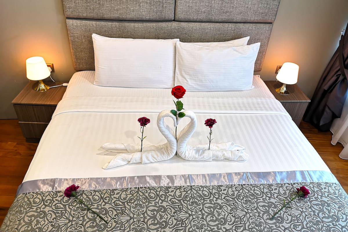 A neatly made bed with white linens, two bedside lamps, and decorative swan pillows with roses.