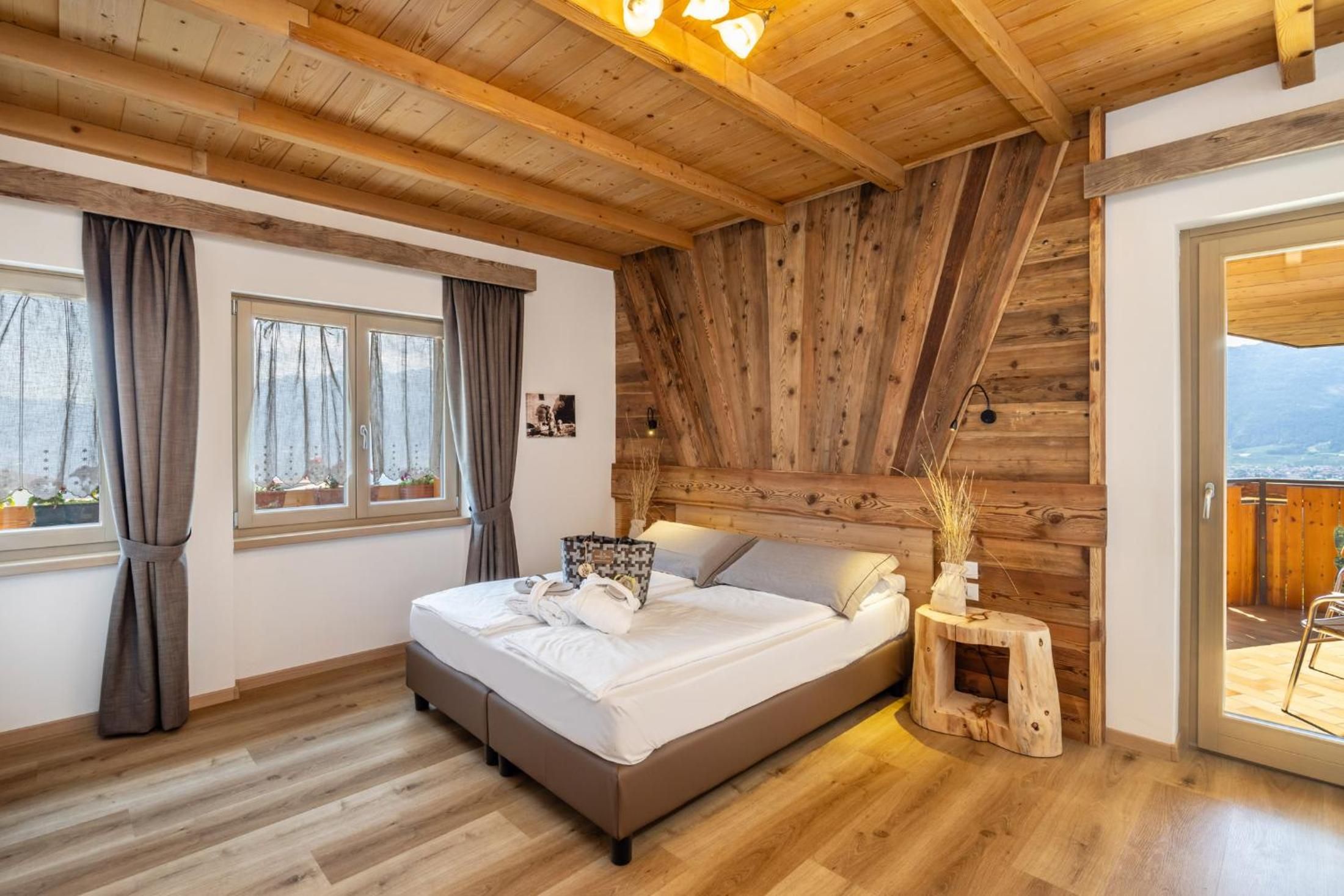 A bedroom with a wooden ceiling, a bed with white bedding, a small wooden side table, and a view through a glass door.