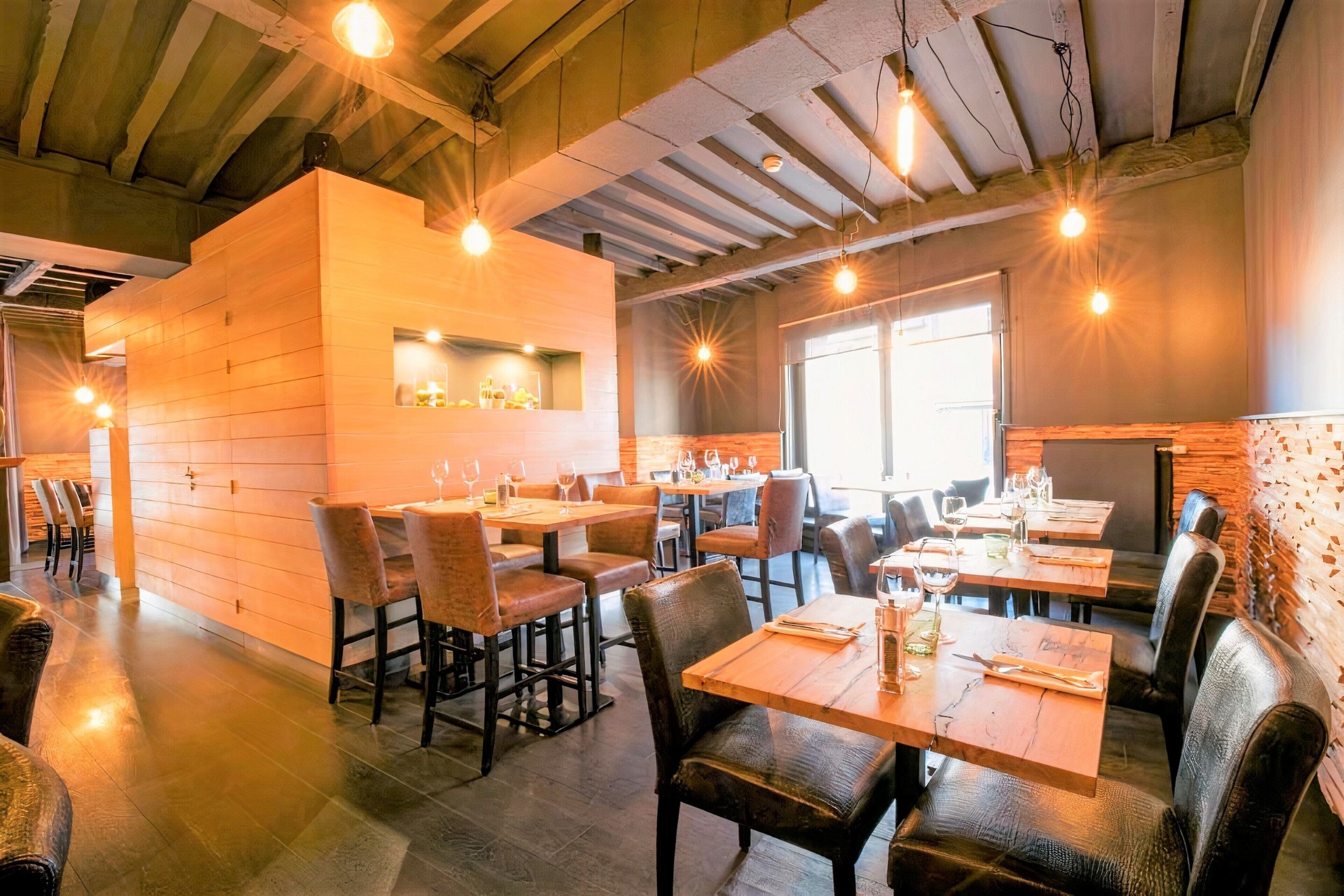 Un restaurante con mesas y sillas de madera, una zona de bar y un amplio ventanal que permite la entrada de luz natural.