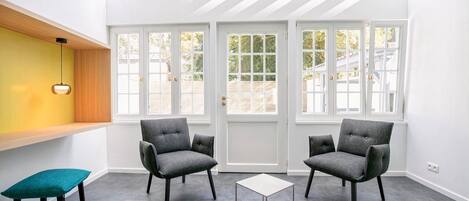 Una habitación luminosa con dos sillones grises, una mesita y un banco. Hay ventanas amplias y un tragaluz.