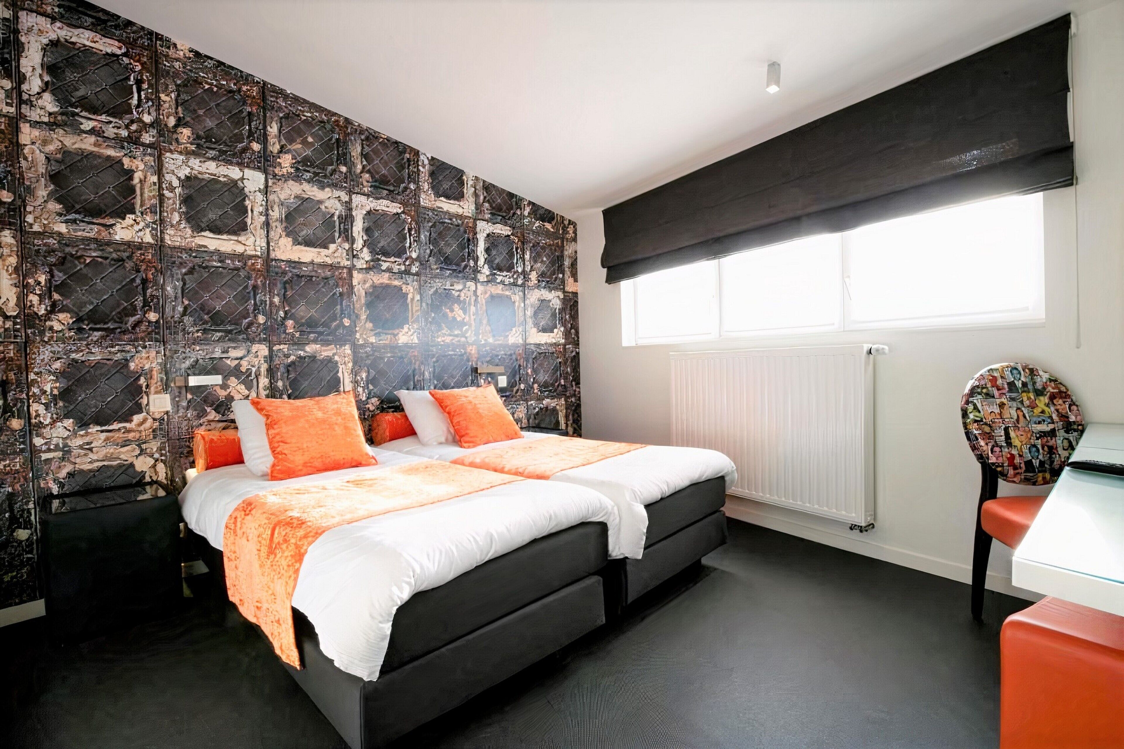 A bedroom with a large bed, a black headboard, a radiator, a window with a black blind, and a chair with a colorful cover.