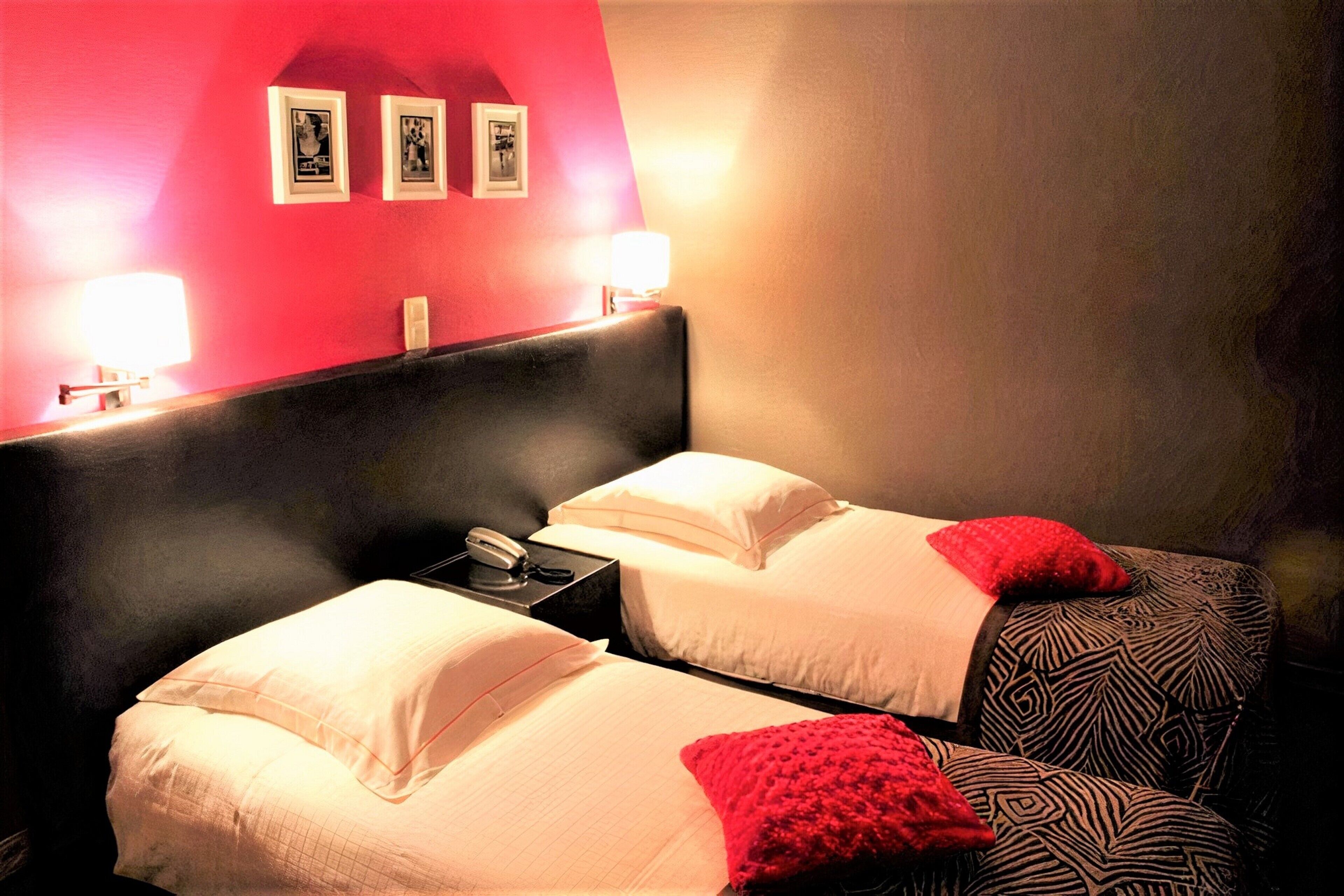 Une chambre d’hôtel avec deux lits, chacun agrémenté de draps blancs et de coussins décoratifs rouges. Des tableaux encadrés ornent le mur au-dessus des lits, et une table de chevet est équipée d’une lampe et d’un téléphone.
