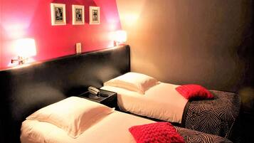 Une chambre d’hôtel avec deux lits, chacun agrémenté de draps blancs et de coussins décoratifs rouges. Des tableaux encadrés ornent le mur au-dessus des lits, et une table de chevet est équipée d’une lampe et d’un téléphone.
