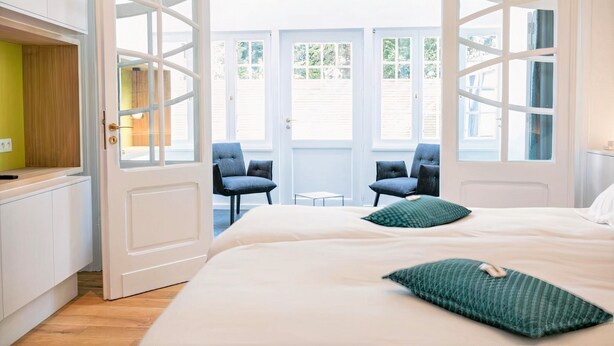 A bedroom with a large bed, two chairs, and a view through open doors. - L'Esprit Sain (Malmedy)