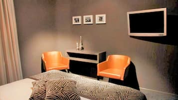 Et hotelværelse med en seng, to orange lænestole, et fjernsyn på væggen og tre billeder i rammer.