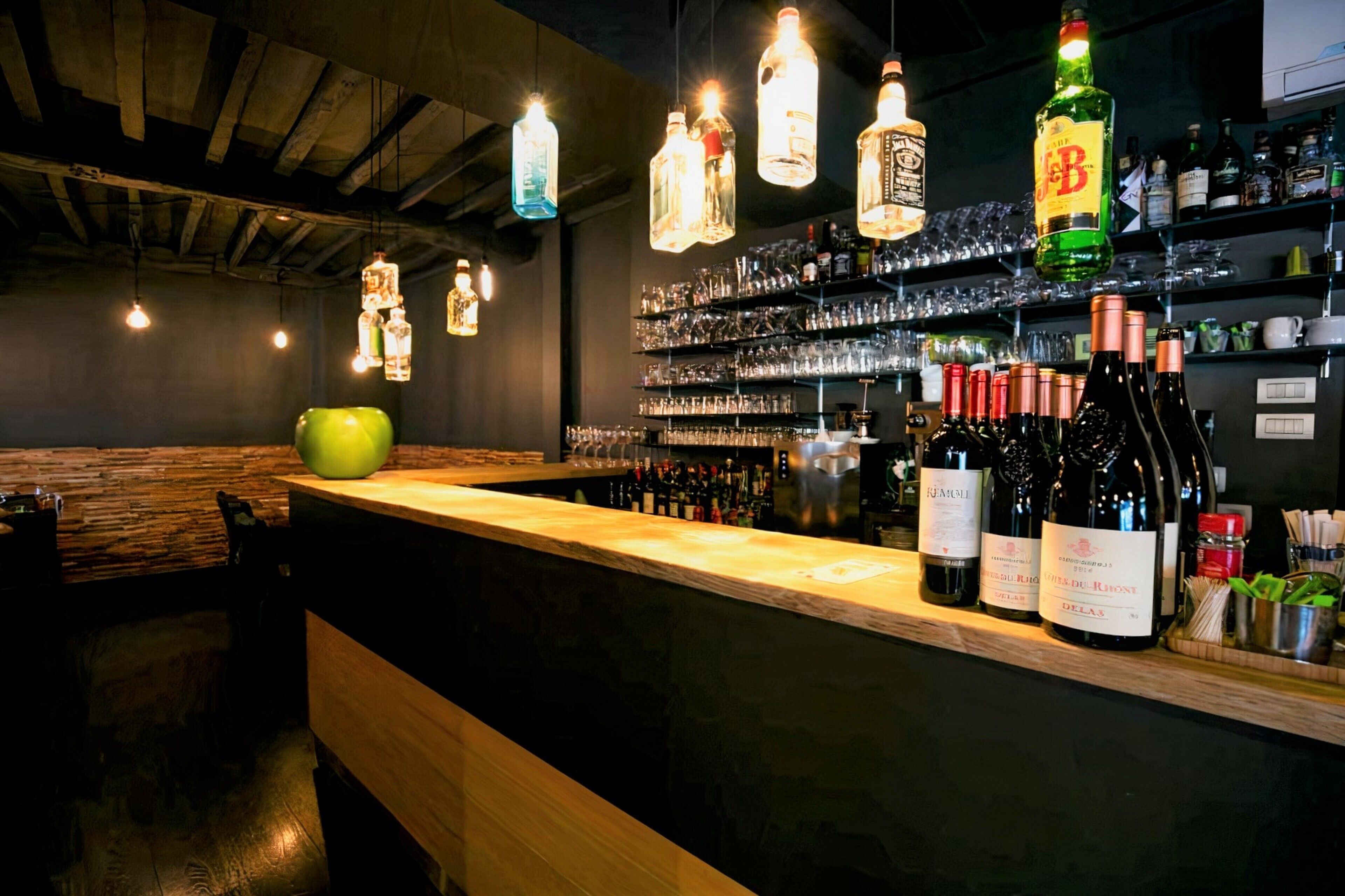 A bar area with wooden counter, hanging light fixtures, and shelves stocked with bottles and glasses.