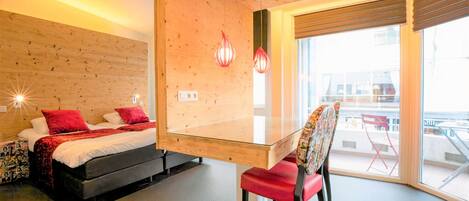 Ein modernes Hotelzimmer mit einer Holzwand, einem Glastisch, einem roten Sessel und einem Bett mit roten Kissen, mit Blick ins Freie.