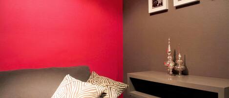 Une pièce avec un mur rouge, un canapé gris agrémenté de coussins à motifs, une console blanche ornée d’objets décoratifs et des tableaux encadrés accrochés au mur.