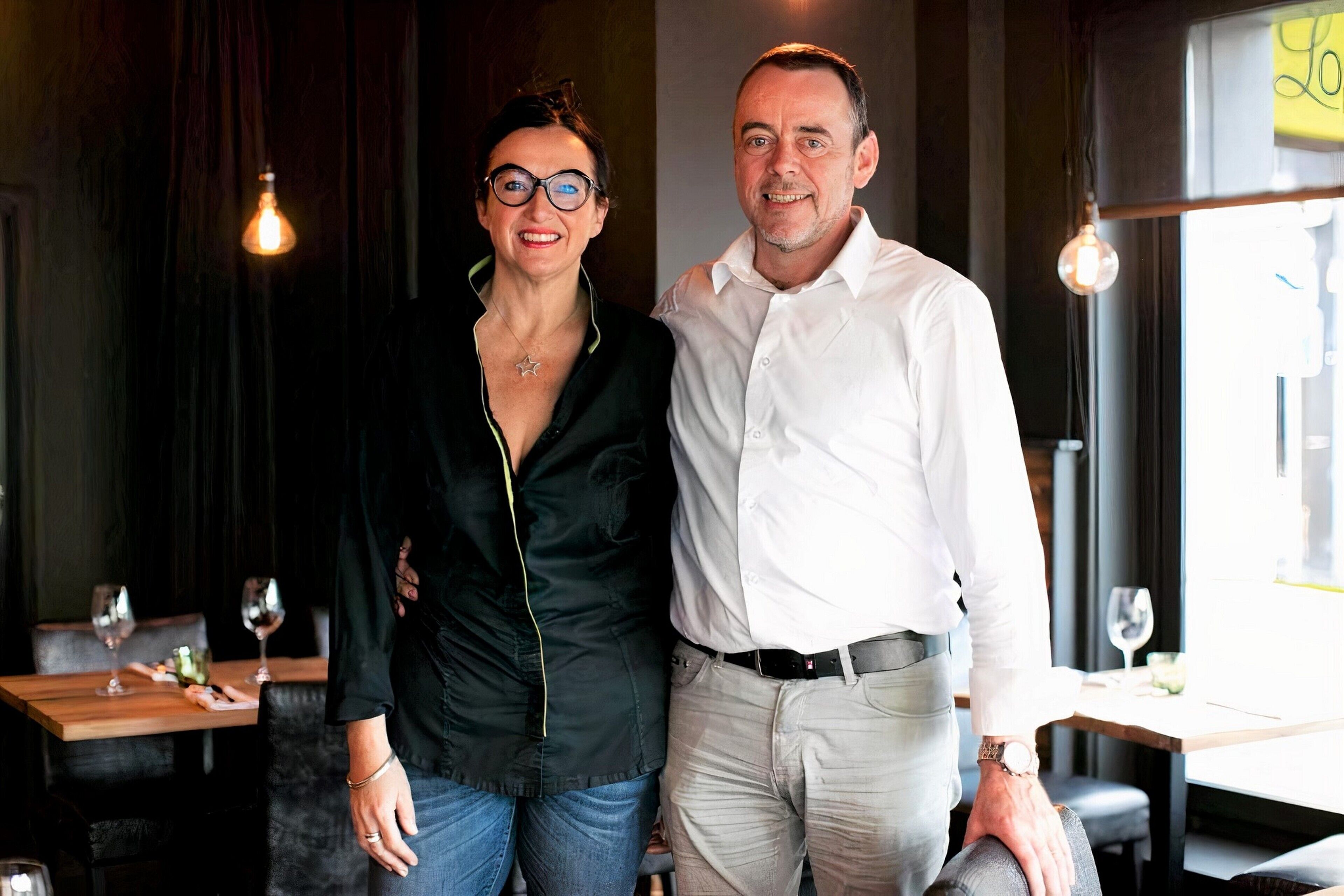 A man and a woman standing in a restaurant with a wooden table and chairs, a hanging light bulb, and a window with a view.