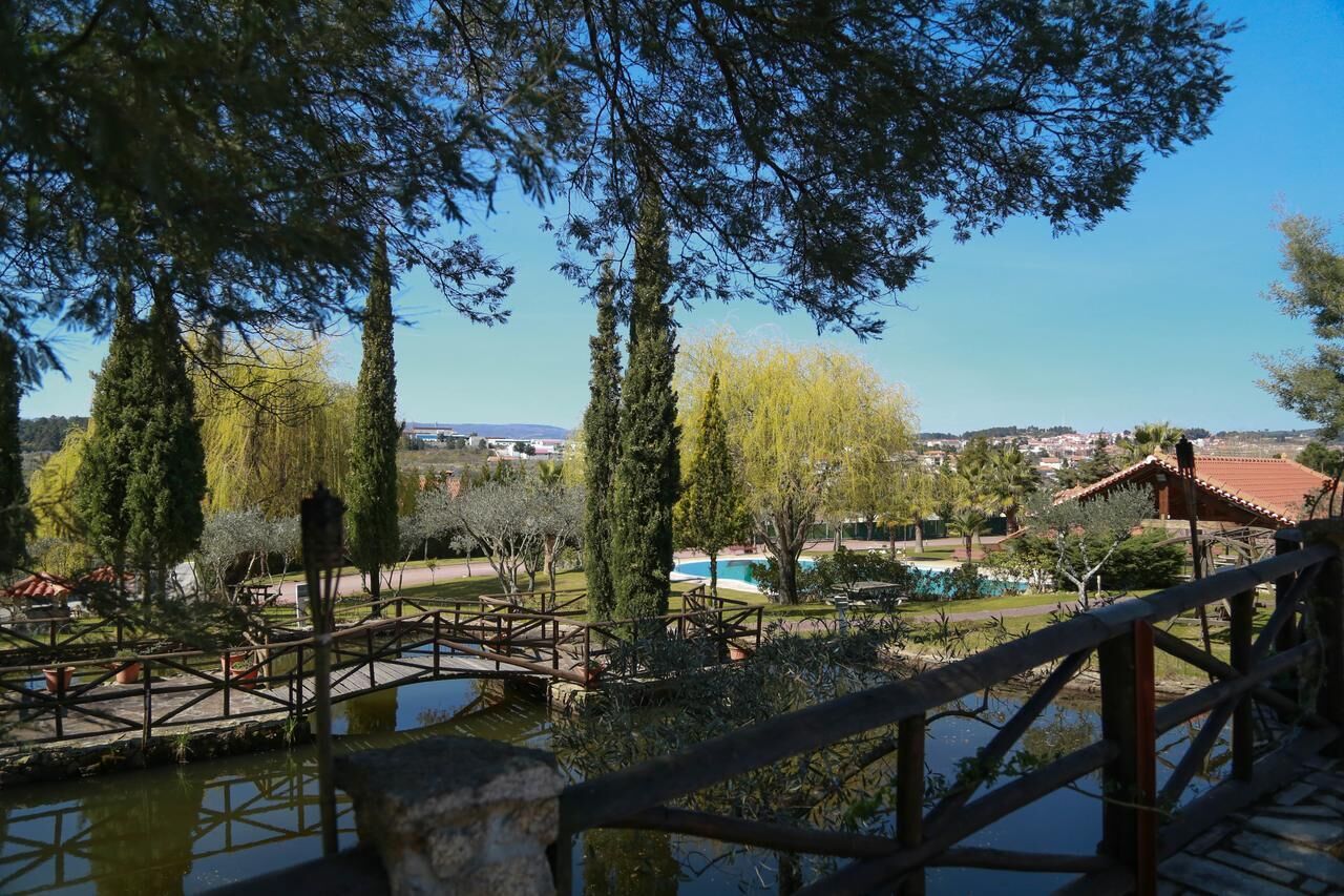 A view of a park with a wooden bridge, a swimming pool, and tall trees.
