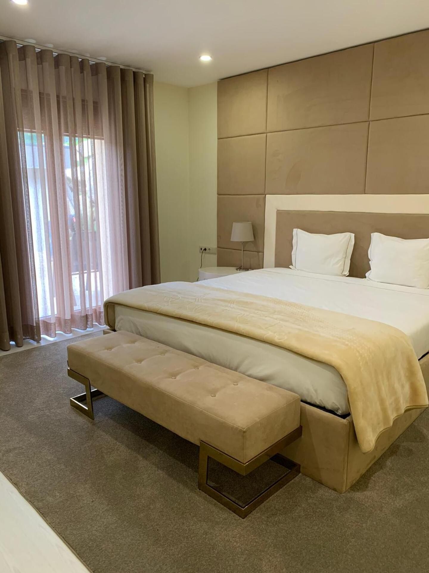 A hotel room with a large bed, a bench, a bedside table with a lamp, and a view through sheer curtains.