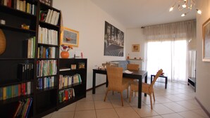 Ein Wohnzimmer mit Bücherregal, Esstisch, Stühlen und einem großen Fenster mit Vorhängen.