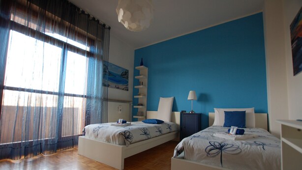 Ein Schlafzimmer mit zwei Betten, einer blauen Akzentwand und einem großen Fenster mit transparenten Vorhängen.