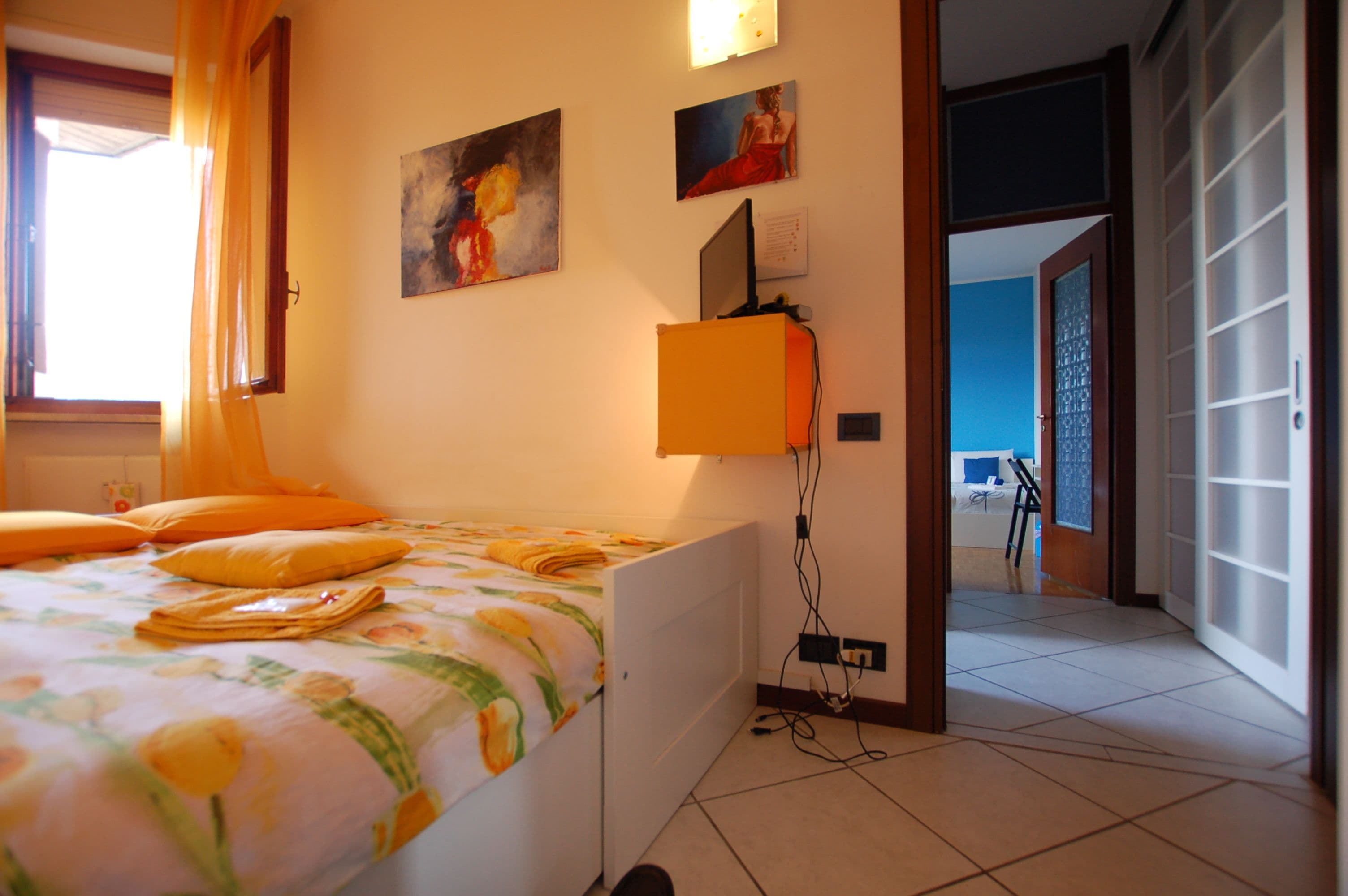 A bedroom with a bed, a desk, and a chair. There are paintings on the wall and a window with curtains.