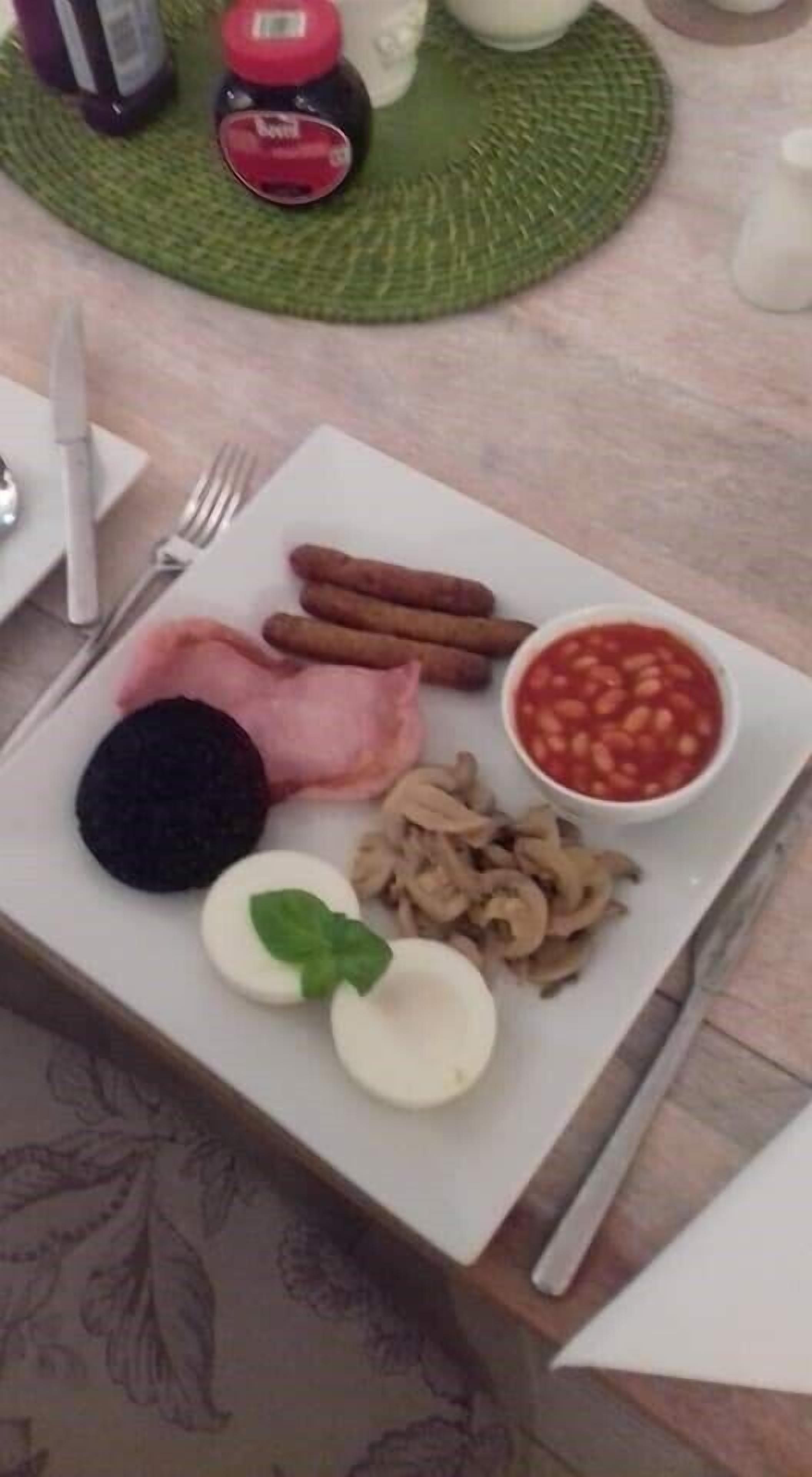 A plate with various food items including beans, mushrooms, a slice of ham, a round black item, and a white round item, possibly cheese, on a table.