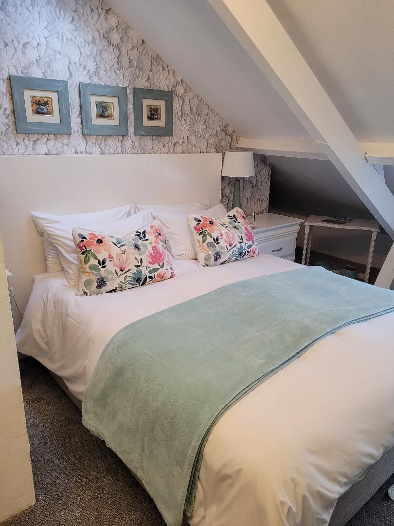 Une chambre à coucher avec un lit, des coussins à motifs floraux, un plaid vert, une table de chevet, une lampe et trois tableaux encadrés accrochés au mur.