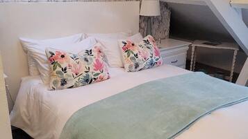 Une chambre à coucher avec un lit, des coussins à motifs floraux, un plaid vert, une table de chevet, une lampe et trois tableaux encadrés accrochés au mur.
