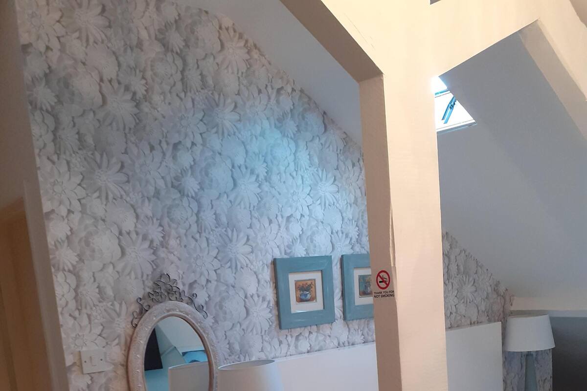 Une chambre avec une commode blanche, un miroir rond, une lampe et un lit. Les murs sont décorés de motifs floraux et on y trouve deux tableaux encadrés.