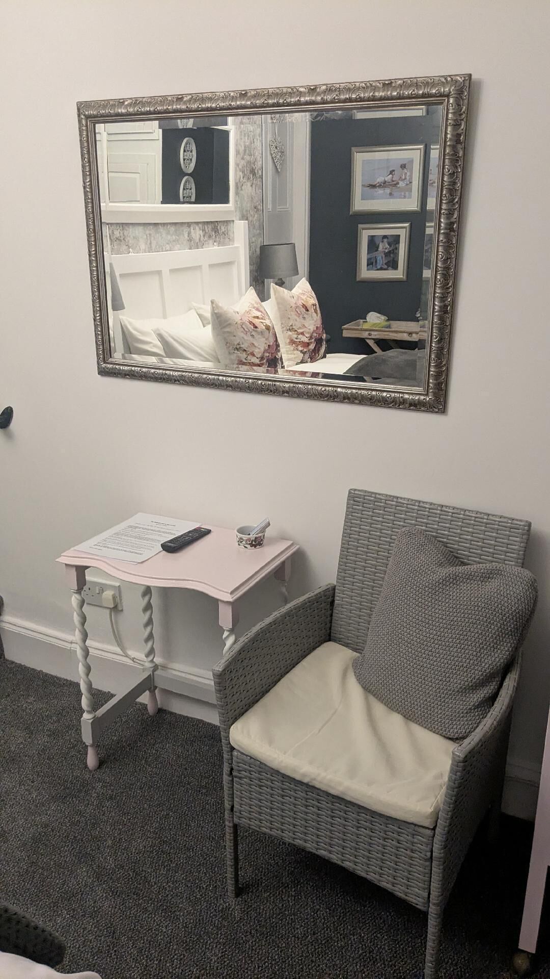 A room with a mirror, a small white table with a remote control, a grey wicker chair, and a carpeted floor.