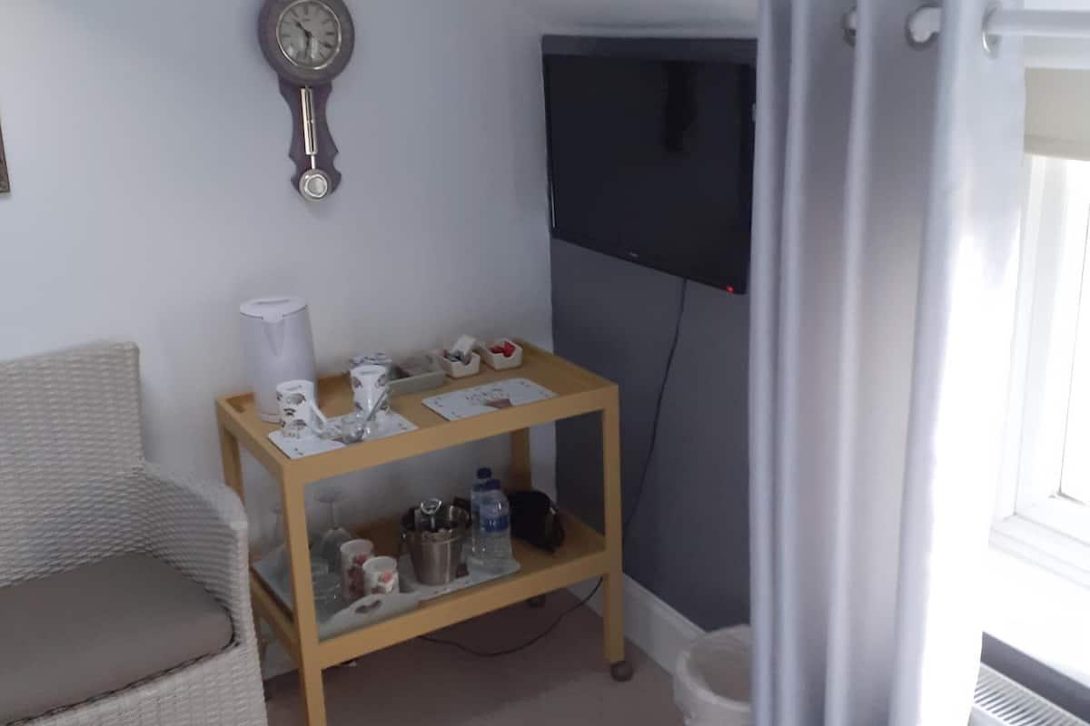 Une petite pièce comprenant un fauteuil en osier, une table d’appoint en bois sur laquelle sont posés divers objets, un téléviseur à écran plat et une fenêtre avec des rideaux.