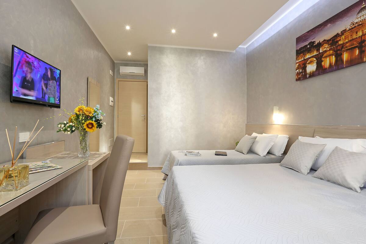 A modern hotel room with a bed, a desk with a vase of flowers, a television, and a painting on the wall.
