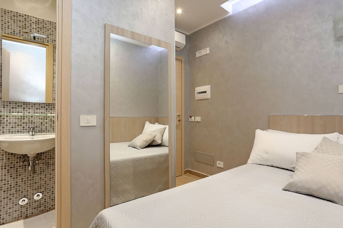 A modern bedroom with a large bed, a bedside mirror, and a bathroom with a sink and mirror.