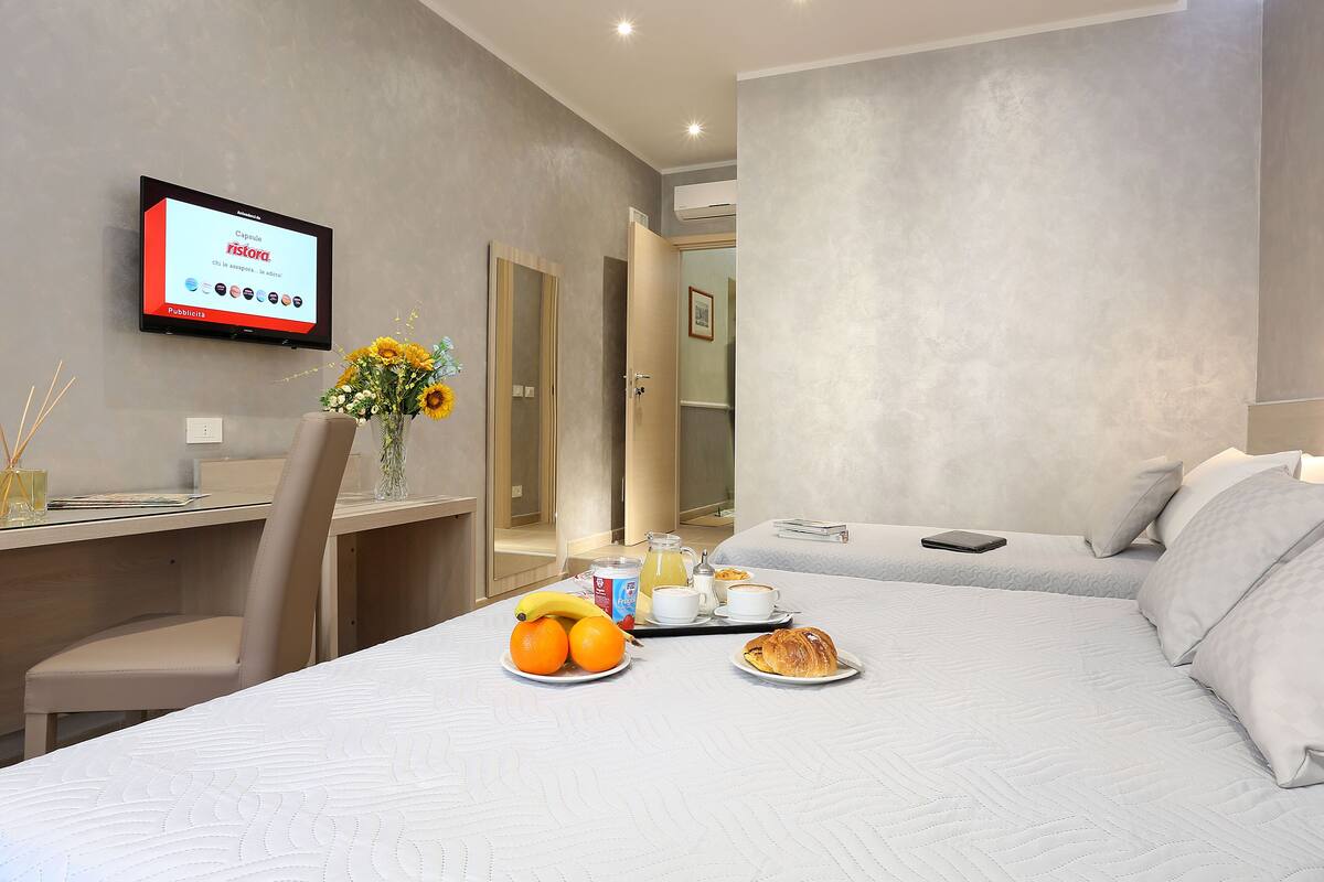 A hotel room with a bed, a desk, a chair, a TV, and a breakfast tray with fruits and pastries.