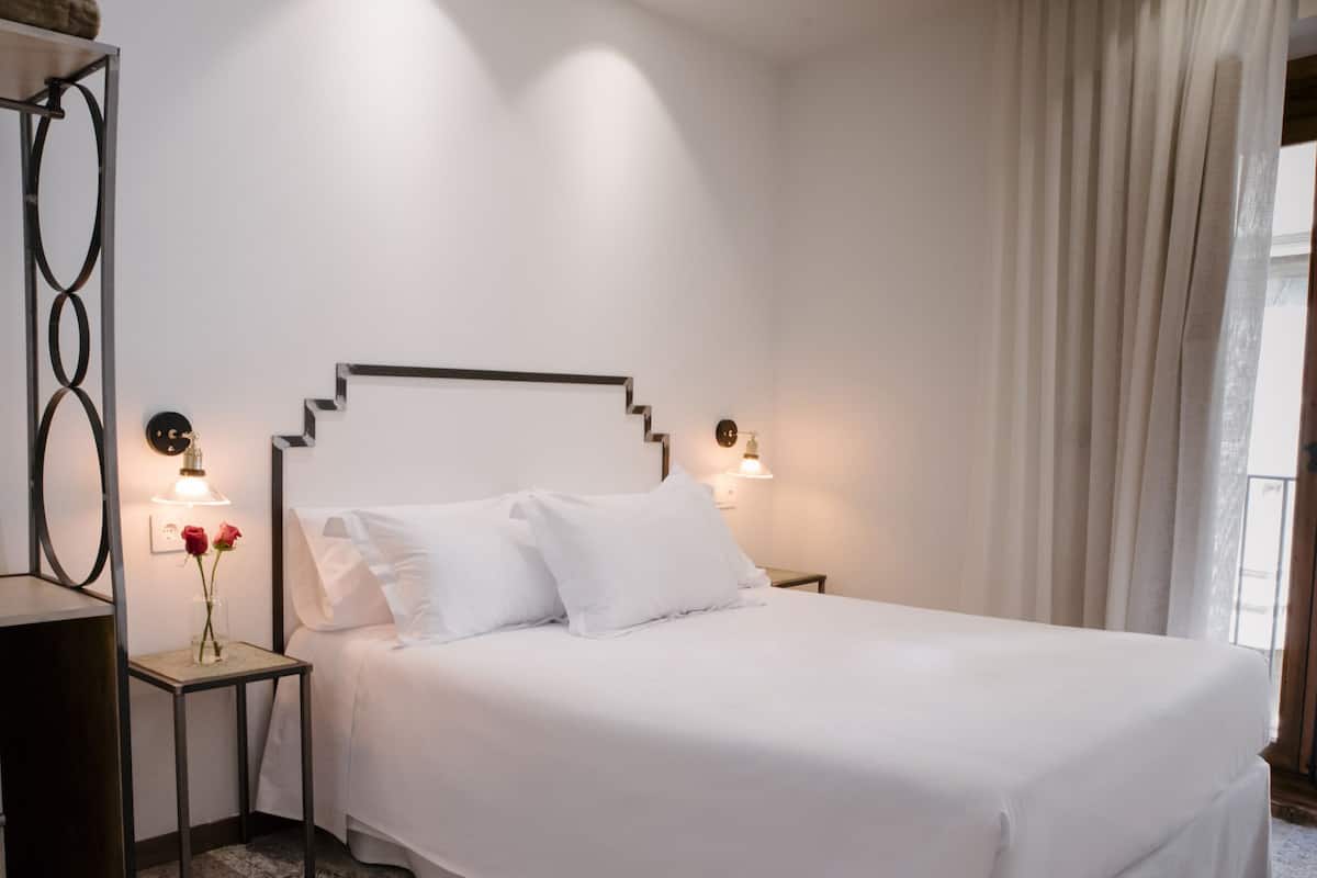 A neatly made bed with white linens in a hotel room. There are two bedside tables with lamps, one on each side of the bed. A large window with curtains is visible on the right side of the room.
