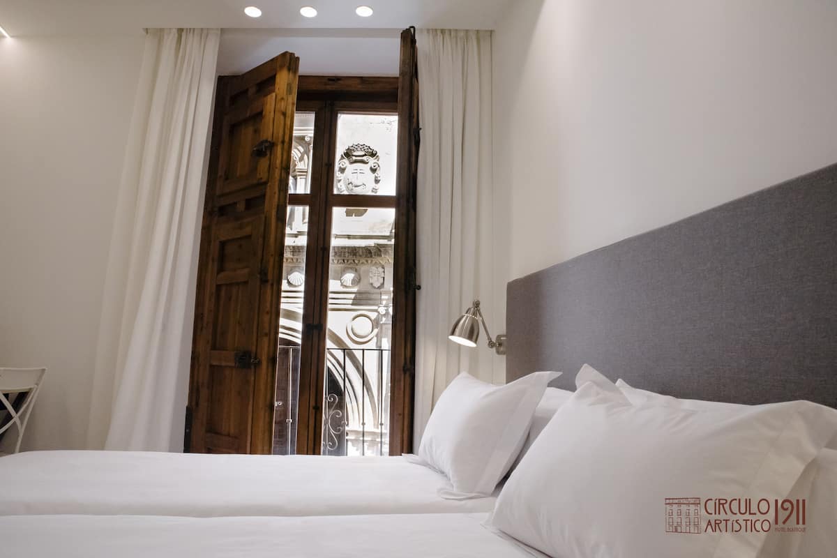 A hotel room with a bed, a wooden door, and a view of a balcony with ornate railings.
