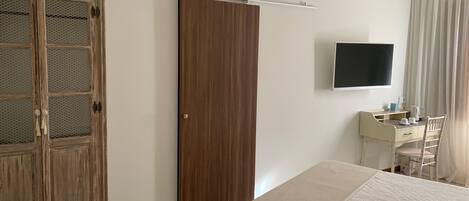 Un dormitorio con cama, armario, escritorio con silla, televisor y ventana con cortinas.