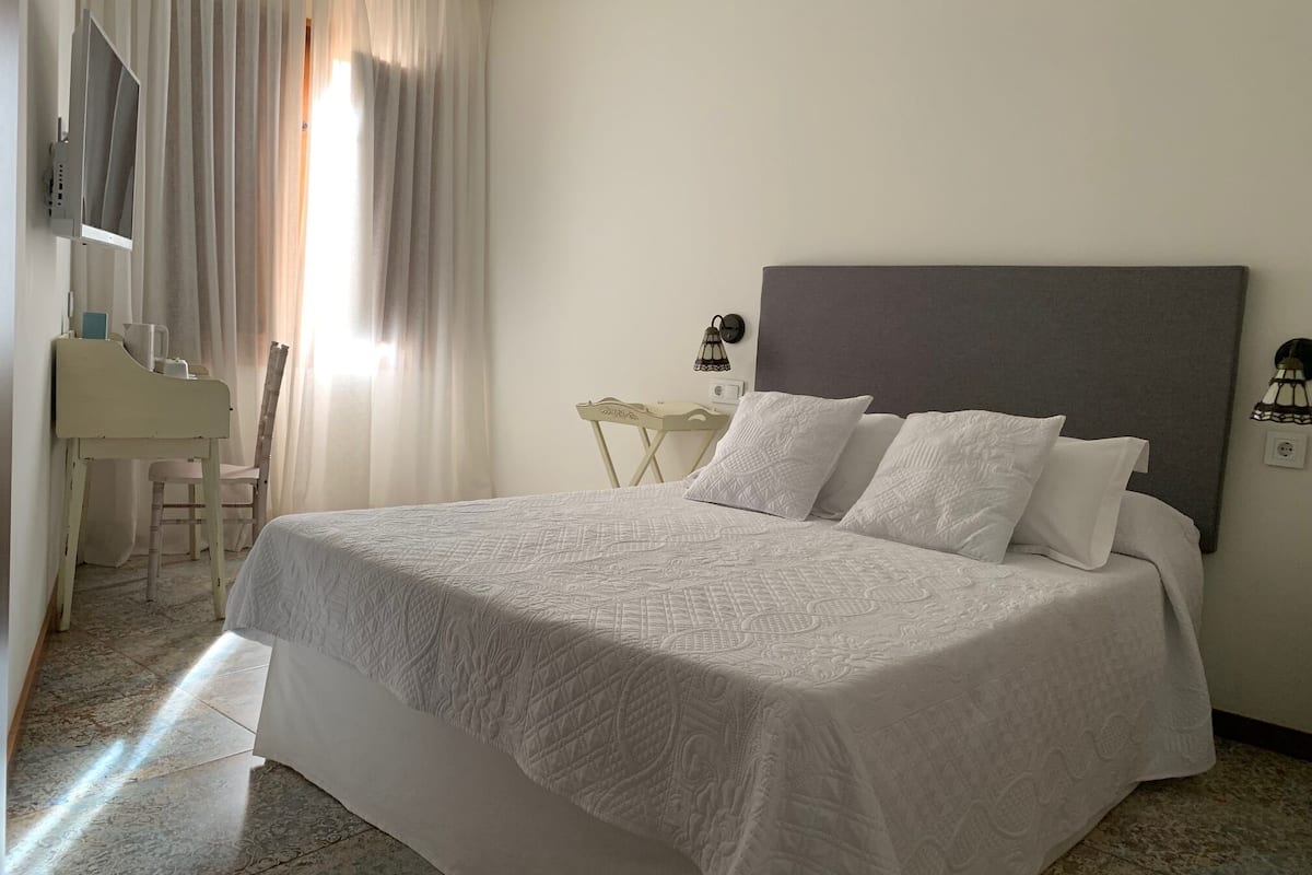 A hotel room with a large bed, a desk with a chair, a lamp, and a window with curtains.