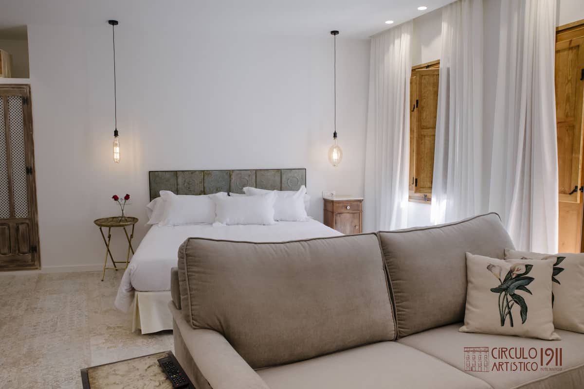 A bedroom with a bed, a wooden nightstand, a round table with a vase of flowers, and a sofa with decorative pillows.