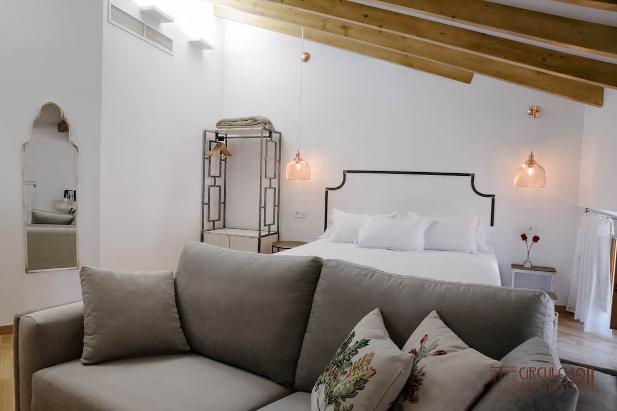 A modern bedroom with a large bed, a grey sofa, and a wooden ceiling.