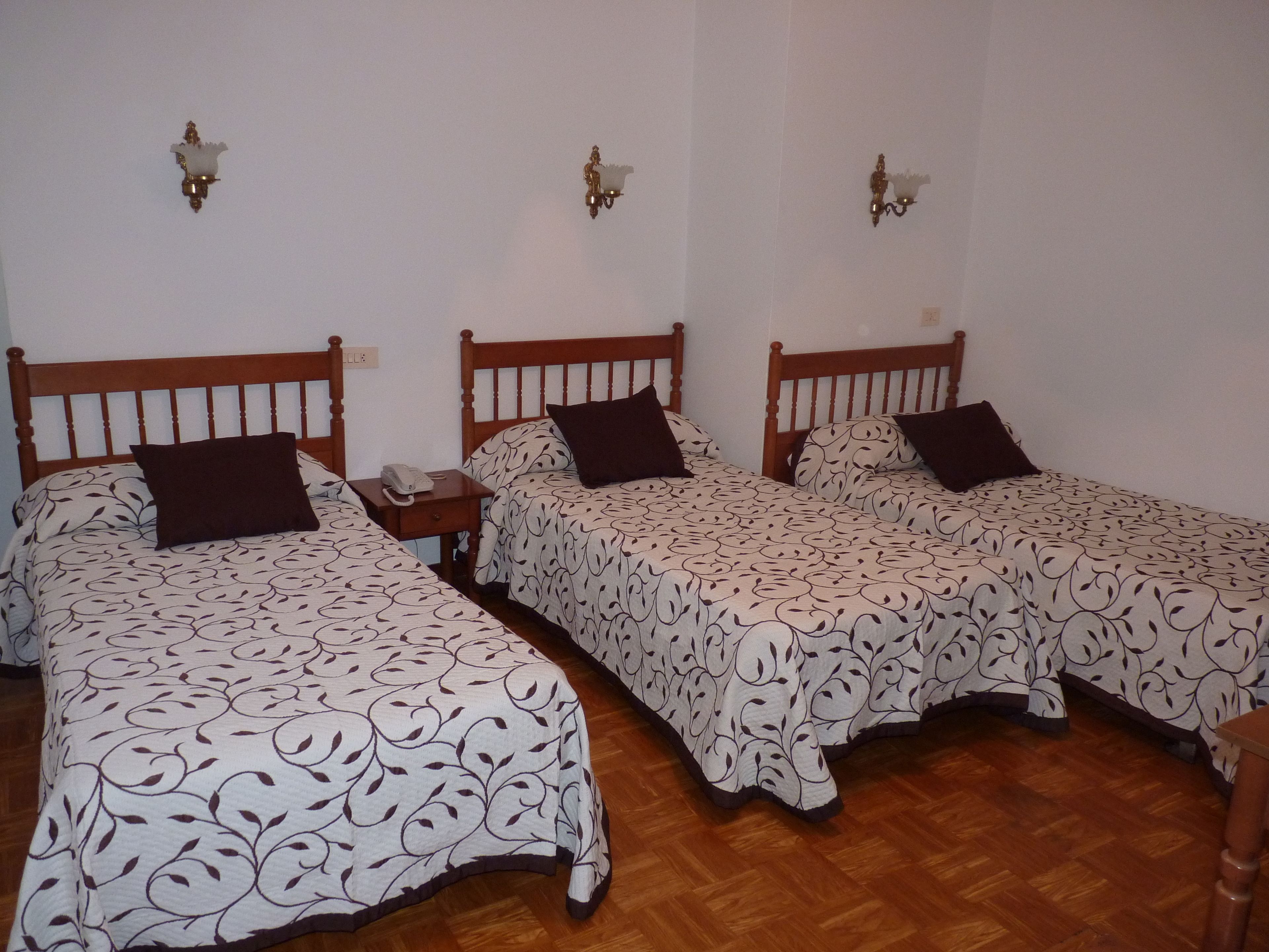 Una habitación con tres camas de madera, cada una con una colcha estampada y un almohadón oscuro. Hay una mesita de noche con un teléfono junto a la cama izquierda.