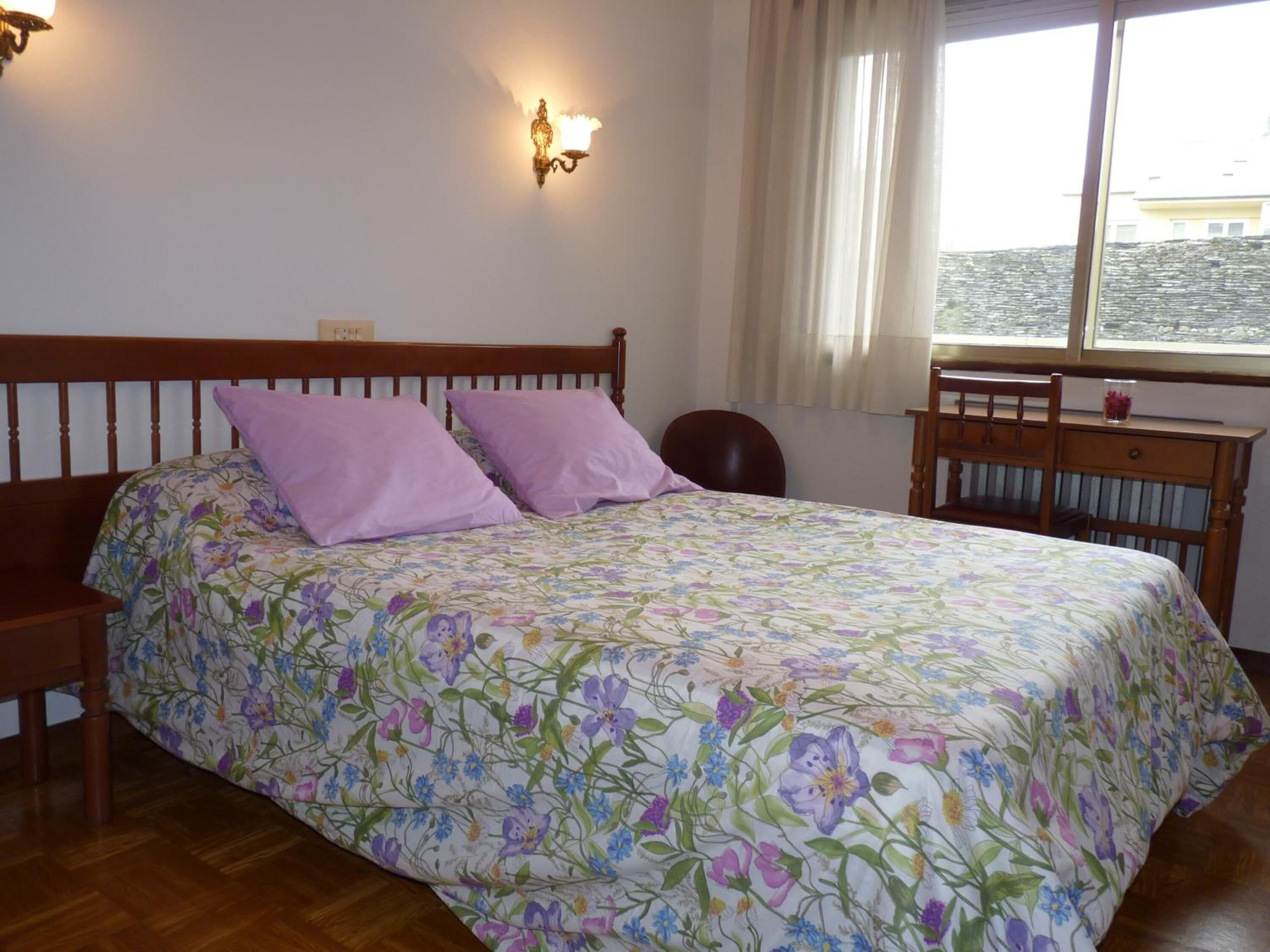 Une chambre avec un lit en bois, une couvre-lit à motifs floraux, deux oreillers, une table de chevet, une chaise et une fenêtre donnant sur des immeubles.